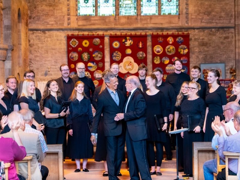 Roderick Williams and Philip Brunelle smiling and wearing dark colored suits shake hands in front of VocalEssence Ensemble Singers wearing black and standing in front of an audience at a church at Three Choirs Festival. Photo Credit: Joseph Wong