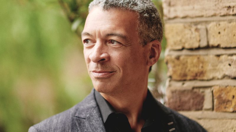 Roderick Williams, wearing a black button down shirt and a dark blazer with a full smile stands in front of a brick wall with trees in the background. Photo Credit: Theo Williams