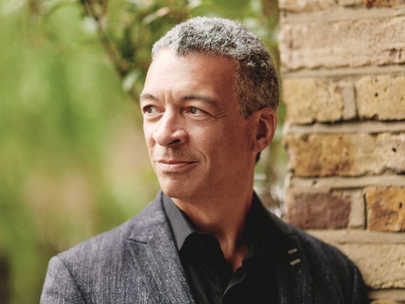 Roderick Williams, wearing a black button down shirt and a dark blazer with a full smile stands in front of a brick wall with trees in the background. Photo Credit: Theo Williams