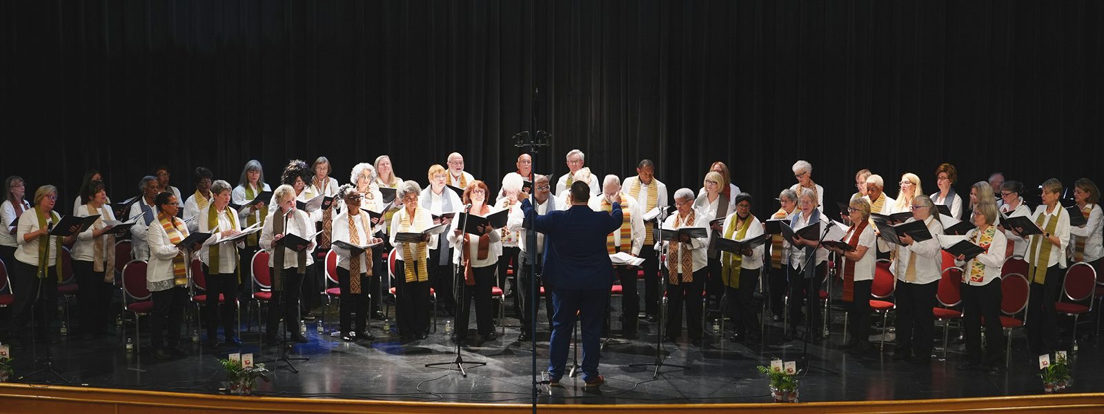 Vintage Voices Singers, conducted by Robert Graham wearing a blue suit, wearing white shirts, black pants, and multi-colored stoles perform on stage in front of a black curtain. Photo credit: Novelli Jurado