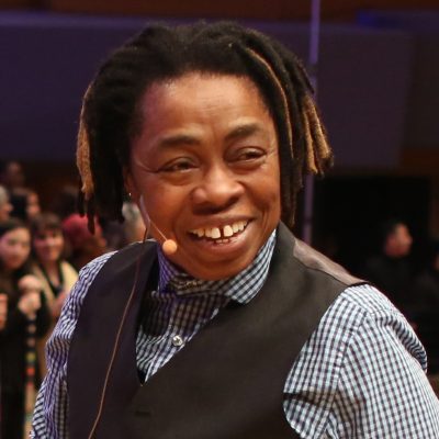 Melanie DeMore, wearing a black vest and bow tie with a checkered white and black shirt smiles. Photo Credit: Kyndell Harkness