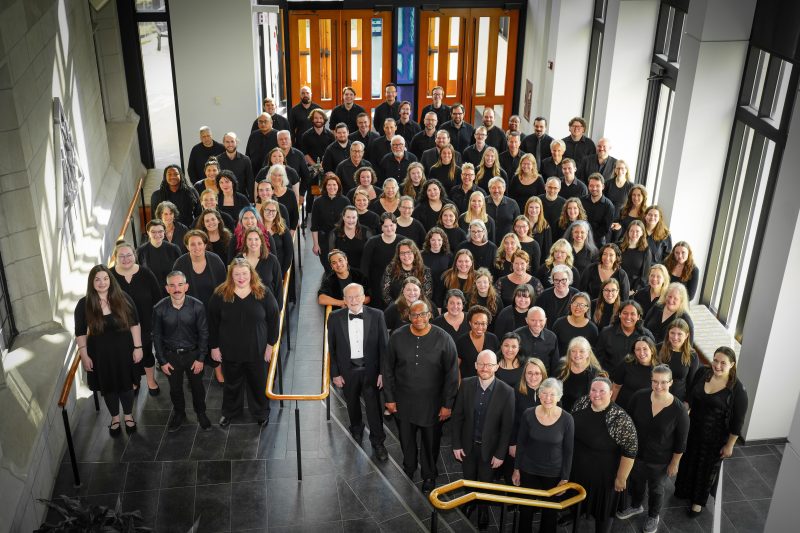 VocalEssence Chorus members wearing black pose for a photo in the lobby of a building with windows and stairs with gold railing. Photo Credit: Novart Media