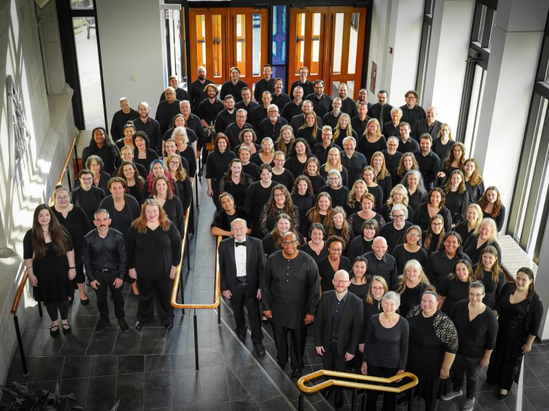 VocalEssence Chorus members wearing black pose for a photo in the lobby of a building with windows and stairs with gold railing. Photo Credit: Novart Media