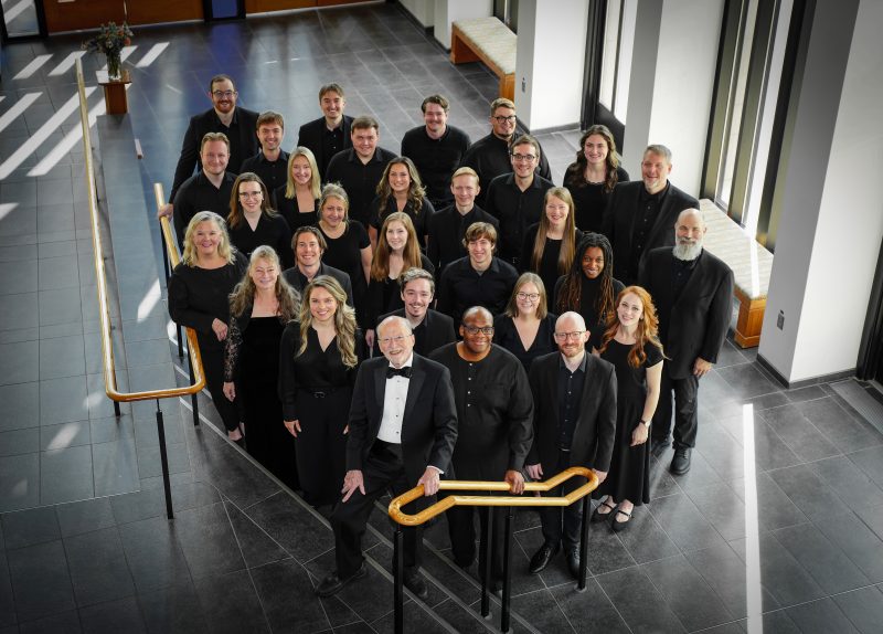 VocalEssence Ensemble Singers wearing black pose for a photo in the lobby of a building with windows and stairs with gold railing. Photo Credit: Novart Media
