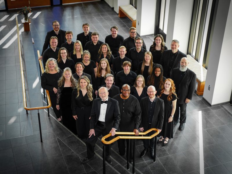 VocalEssence Ensemble Singers wearing black pose for a photo in the lobby of a building with windows and stairs with gold railing. Photo Credit: Novart Media