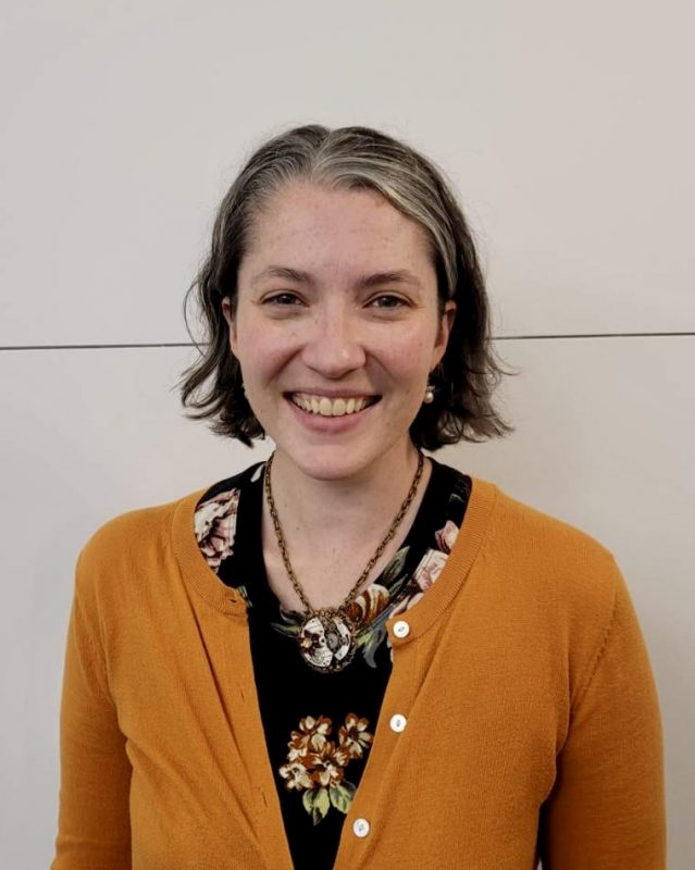 A person with a bob haircut wearing a floral shirt and a goldenrod cardigan.