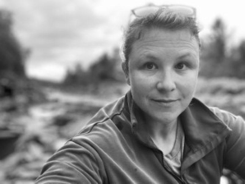 Black and white image of a person with glasses on the top of her head wearing a zip up jacket in front of a nature background. 