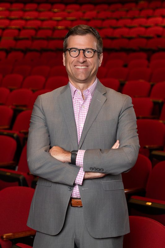 Person wearing glasses a gray suit and pink and white button down shirt with arms folded standing in front of red chairs in an auditorium.