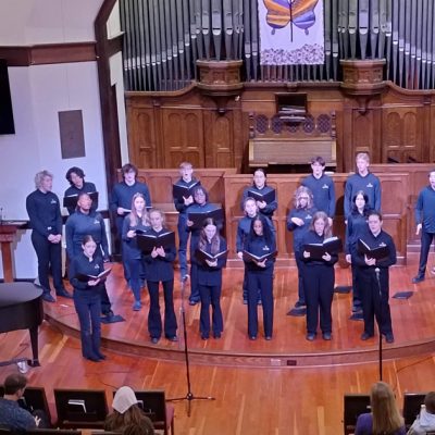 VocalEssence Singers Of This Age, wearing blue shirts and black pants holding black folders sing in the sanctuary of a church. Photo credit: Rhiannon Fiskradatz
