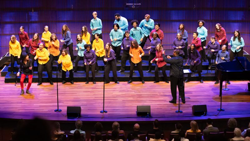 Image of singers on stage wearing multicolored shirts and black pants dancing and singing. Photo Credit: Anna Min