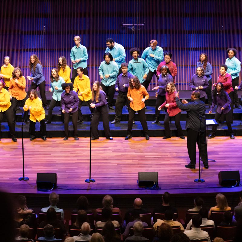 Image of singers on stage wearing multicolored shirts and black pants dancing and singing. Photo Credit: Anna Min