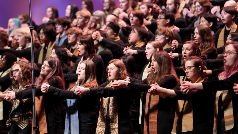 VocalEssence singers wearing black with multicolored stoles perform while holding their arms forward on stage for the annual WITNESS concert. Photo Credit: Kyndell Harkness