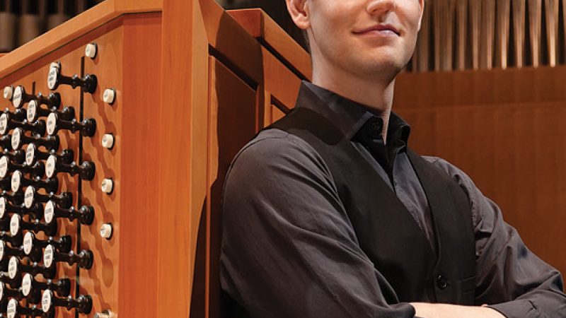 Greg Zelek, wearing a black shirt and vest with hands folded poses in front of an organ console and pipes. Photo Credit: Peter Rodgers