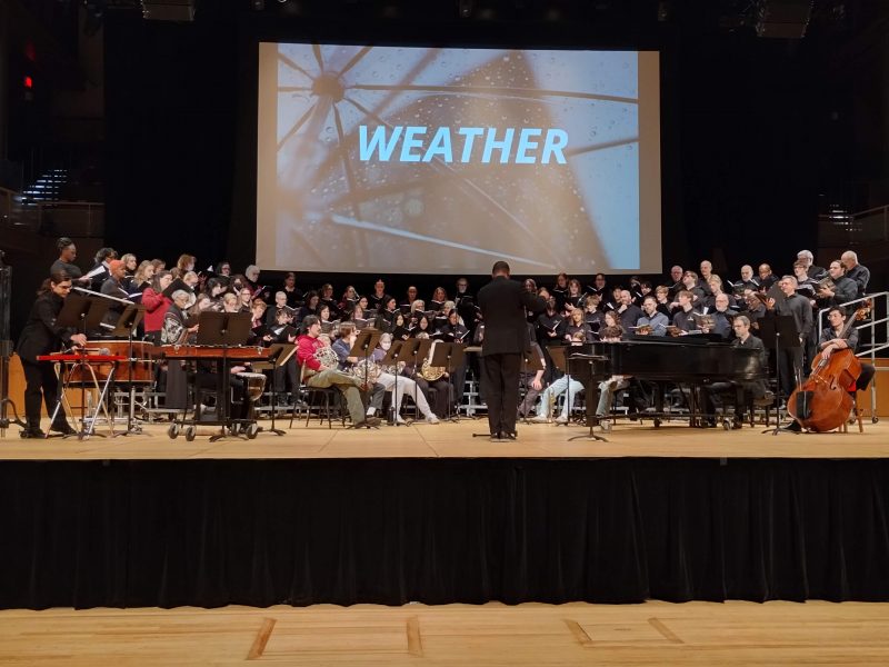 Singers on stage on risers in front of a screen with the word, "Weather" on it.