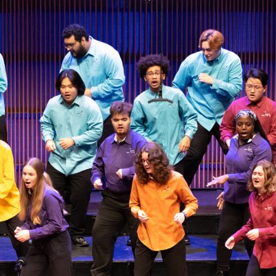 Members of VocalEssence Singers Of This Age wearing multicolored shirts and black pants singing and dancing on risers. Photo credit: Anna Min