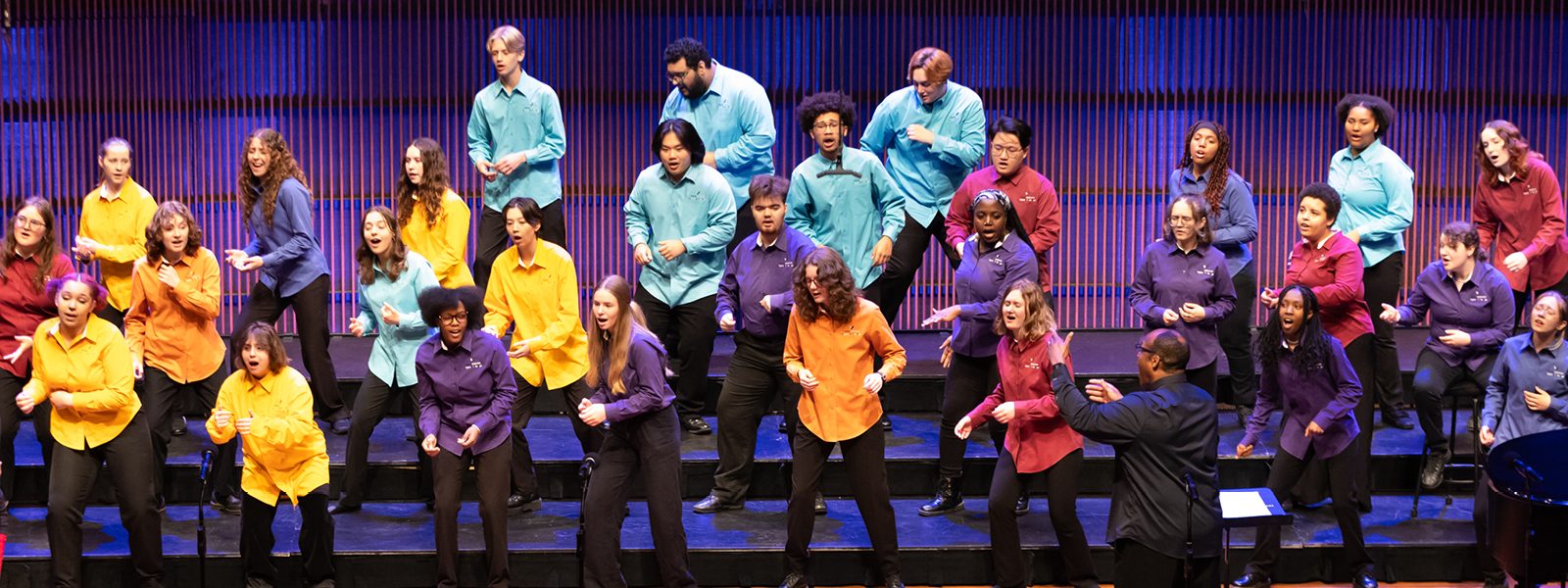 Members of VocalEssence Singers Of This Age wearing multicolored shirts and black pants singing and dancing on risers. Photo credit: Anna Min