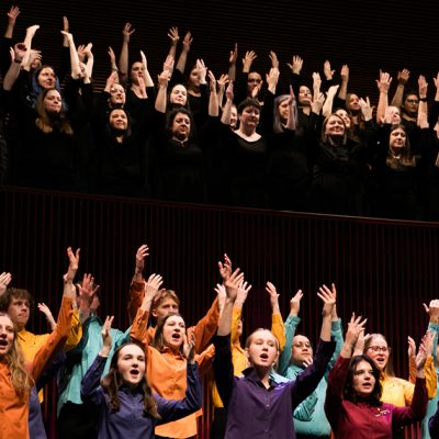 VocalEssence Singers performing, one group of singers is in the balcony, wearing black with their hands raised up and another group is on the stage wearing different colored shirts with their hands raised up. Photo credit: BelleAmiss Photography