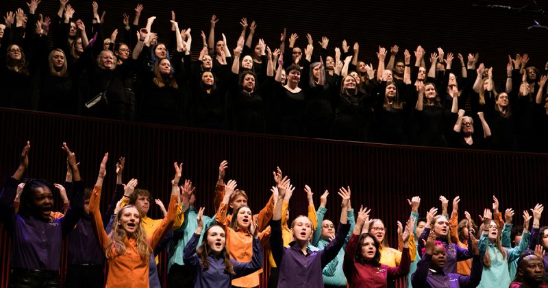 VocalEssence Singers performing, one group of singers is in the balcony, wearing black with their hands raised up and another group is on the stage wearing different colored shirts with their hands raised up. Photo credit: BelleAmiss Photography