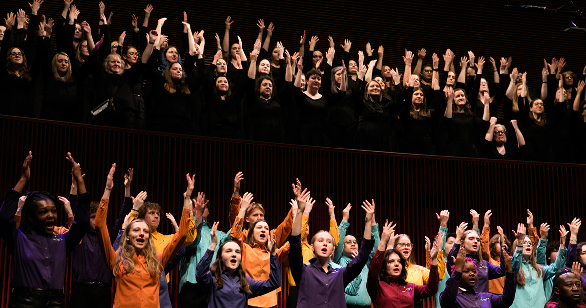 VocalEssence Singers performing, one group of singers is in the balcony, wearing black with their hands raised up and another group is on the stage wearing different colored shirts with their hands raised up. Photo credit: BelleAmiss Photography