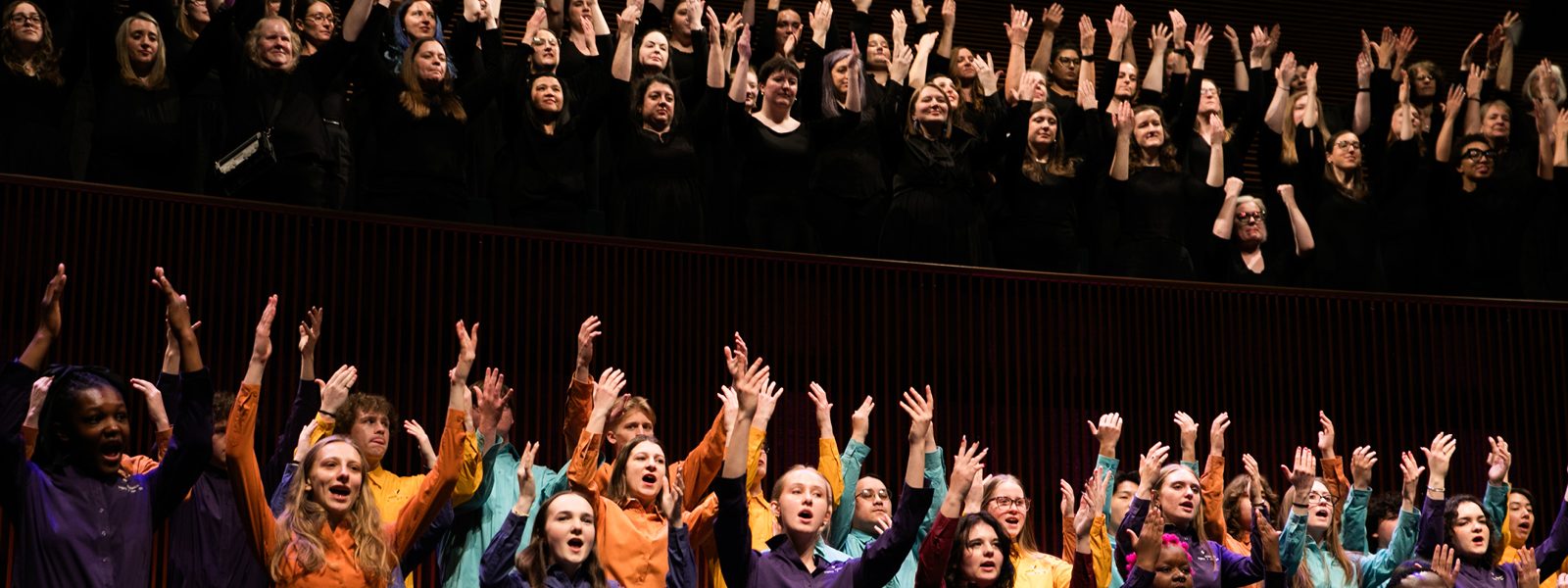 VocalEssence Singers performing, one group of singers is in the balcony, wearing black with their hands raised up and another group is on the stage wearing different colored shirts with their hands raised up. Photo credit: BelleAmiss Photography
