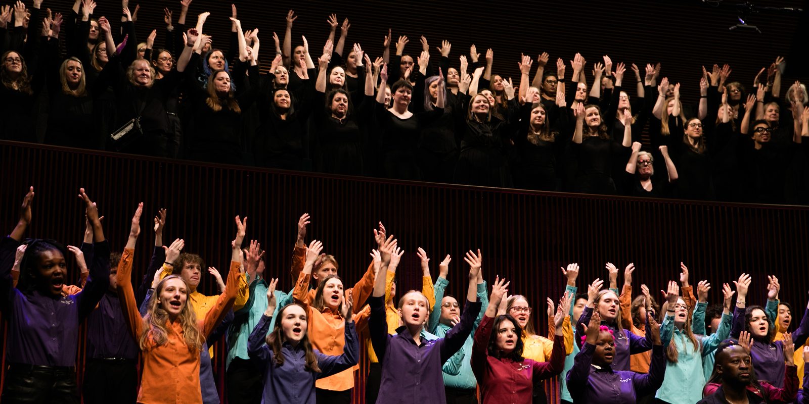 VocalEssence Singers performing, one group of singers is in the balcony, wearing black with their hands raised up and another group is on the stage wearing different colored shirts with their hands raised up. Photo credit: BelleAmiss Photography