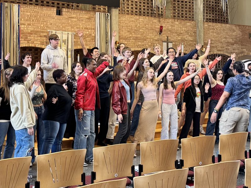 Members of Vox A Cappella Band and VocalEssence Singers Of This Age dancing with their arms outstretched and singing in a church sanctuary with rows of chairs in front of them. Photo Credit: Emma Jirele