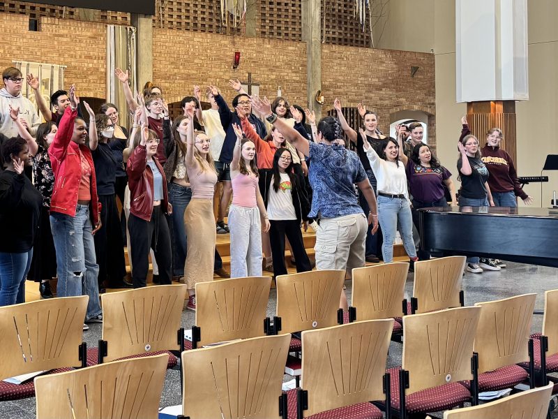 Julio Morales at the grand piano in front of members of Vox A Cappella Band and VocalEssence Singers Of This Age dancing with their hands outstretched and singing in a church sanctuary with rows of chairs in front of them. Photo Credit: Emma Jirele