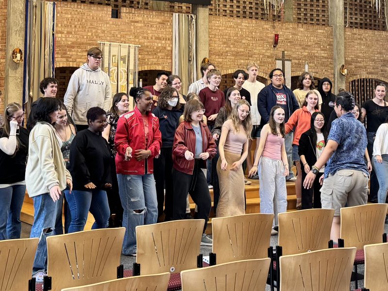 Members of Vox A Cappella Band and VocalEssence Singers Of This Age dancing and singing in a church sanctuary with rows of chairs in front of them. Photo Credit: Emma Jirele