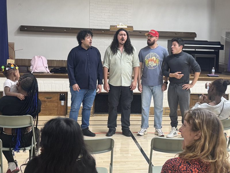Members of Vox A Cappella Band standing in front of a stage and performing for students and their children sitting in folding chairs. Photo credit: Emma Jirele