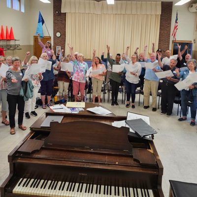 Image of singers with their hands up around a piano. Photo Credit: Robert Graham