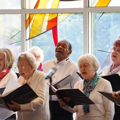 Adult Vintage Voices members wearing white shirts and different colored neckwear holding black folders and singing in front of a stained glass window. Photo Credit: Novart Media.