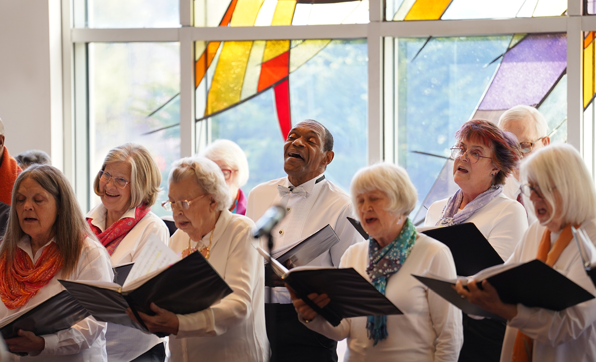 Adult Vintage Voices members wearing white shirts and different colored neckwear holding black folders and singing in front of a stained glass window. Photo Credit: Novart Media.