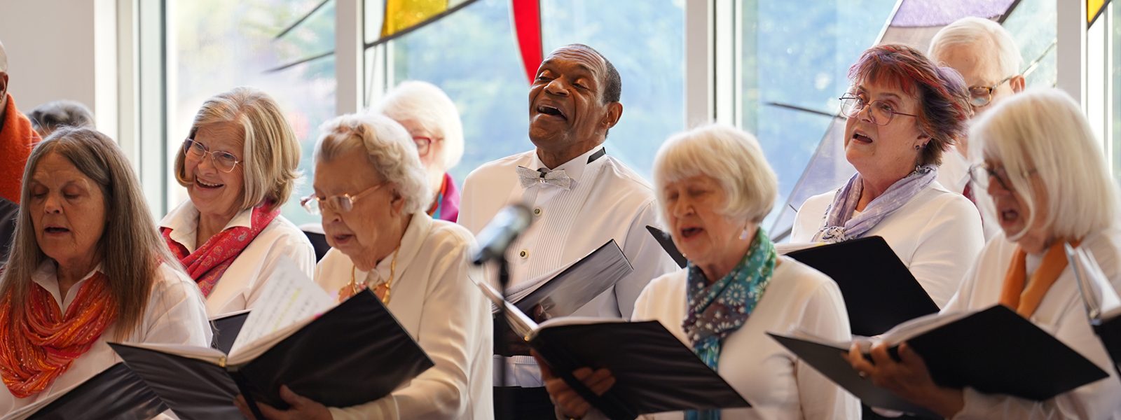 Adult Vintage Voices members wearing white shirts and different colored neckwear holding black folders and singing in front of a stained glass window. Photo Credit: Novart Media.