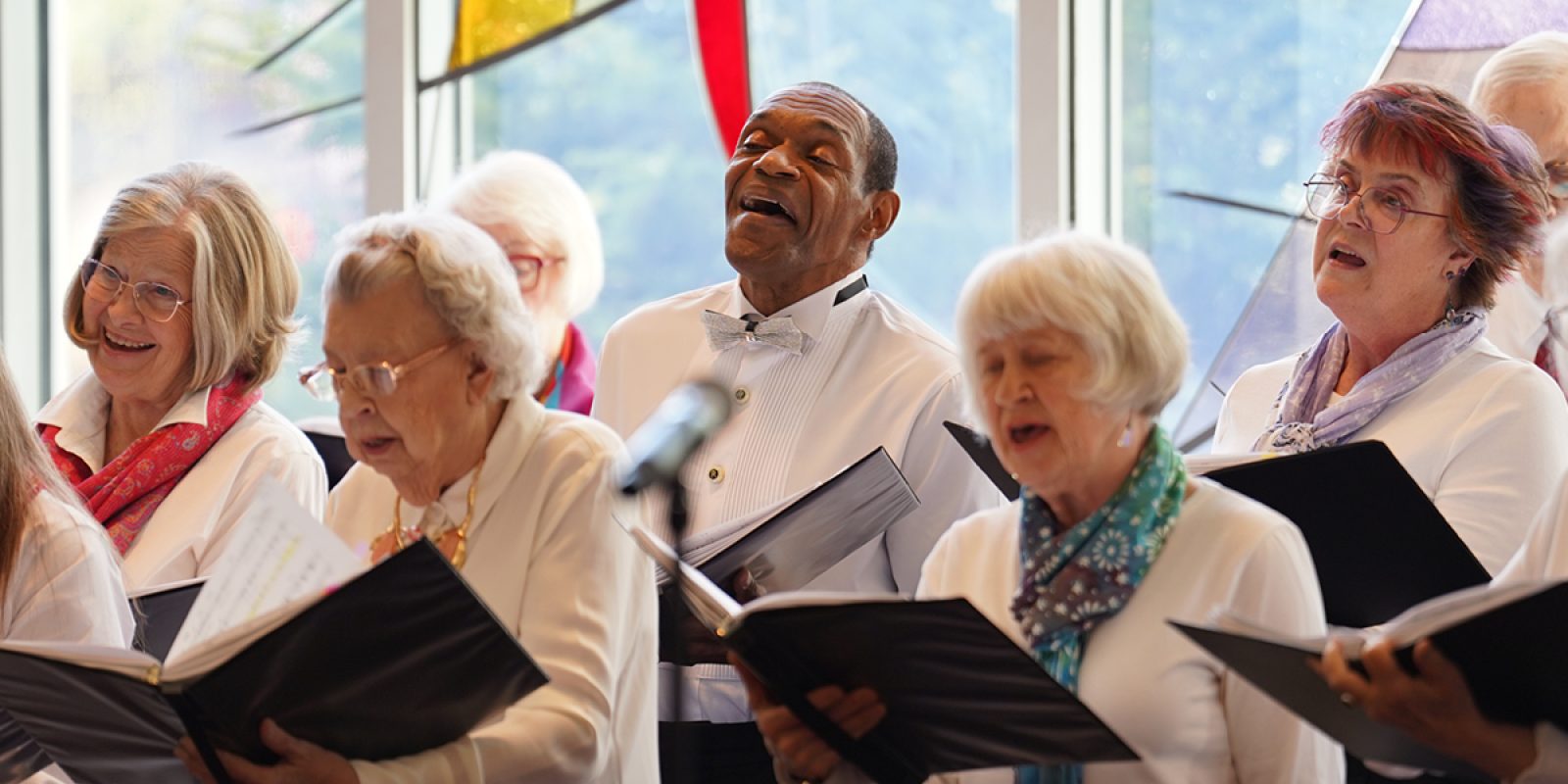 Adult Vintage Voices members wearing white shirts and different colored neckwear holding black folders and singing in front of a stained glass window. Photo Credit: Novart Media.