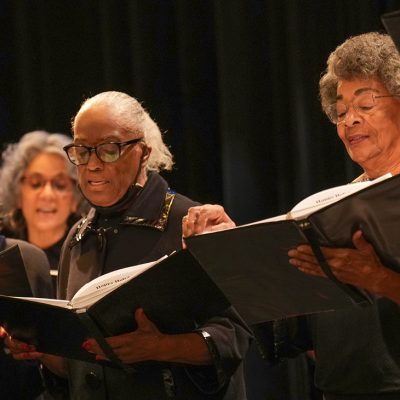 Adult Vintage Voices members wearing all black holding black folders and singing in front of a black curtain. Photo Credit: Novart Media.