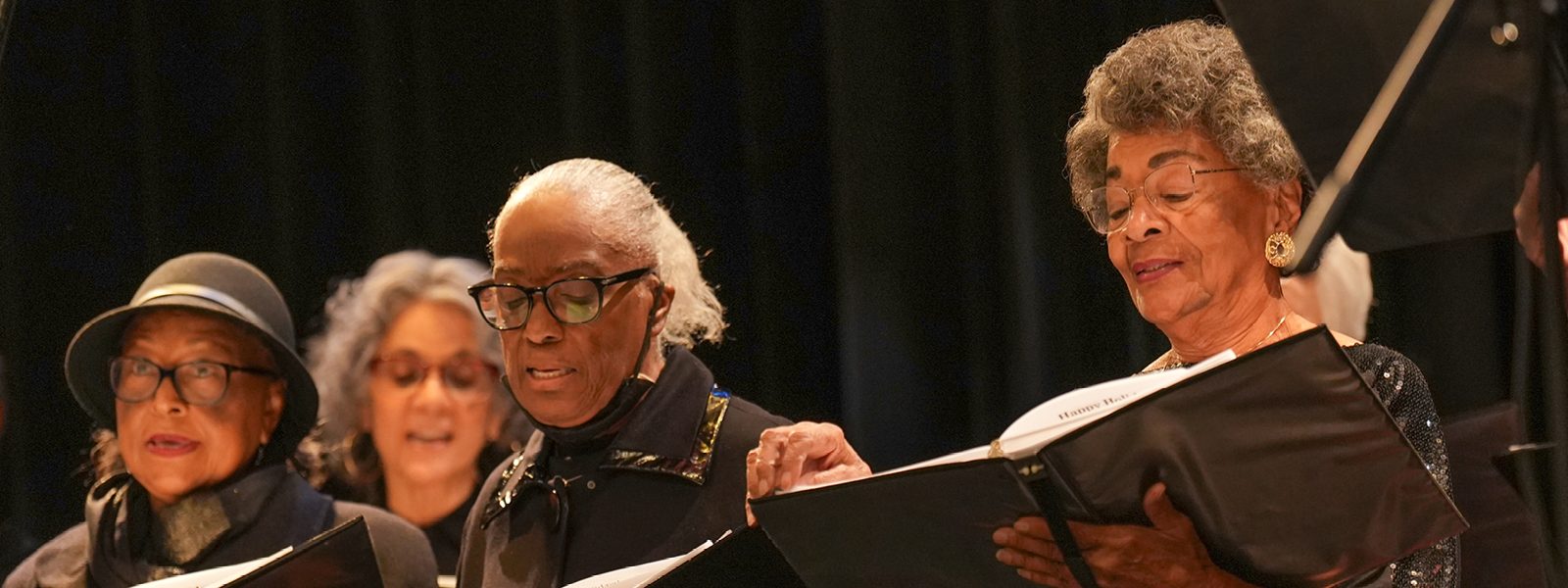 Adult Vintage Voices members wearing all black holding black folders and singing in front of a black curtain. Photo Credit: Novart Media.