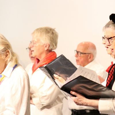 Adult Vintage Voices members wearing white shirts and different colored scarves holding black folders and singing. Photo Credit: Tonia Swennes.
