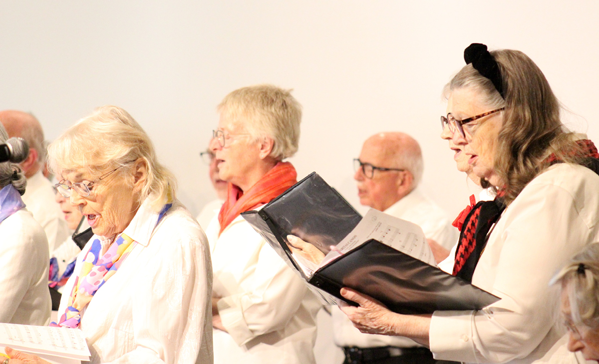 Adult Vintage Voices members wearing white shirts and different colored scarves holding black folders and singing. Photo Credit: Tonia Swennes.