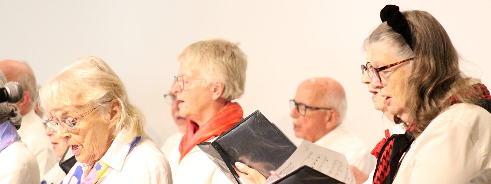 Adult Vintage Voices members wearing white shirts and different colored scarves holding black folders and singing. Photo Credit: Tonia Swennes.