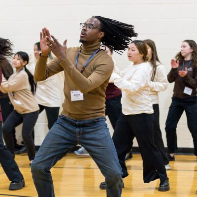 Teenage participants in the Urban Choir Festival wearing long sleeved shirts of different colors and black pants while clapping and dancing led by an instructor with facial hair and glasses wearing a tan turtleneck and jeans clapping and dancing. Photo Credit: Kara Salava Photography.
