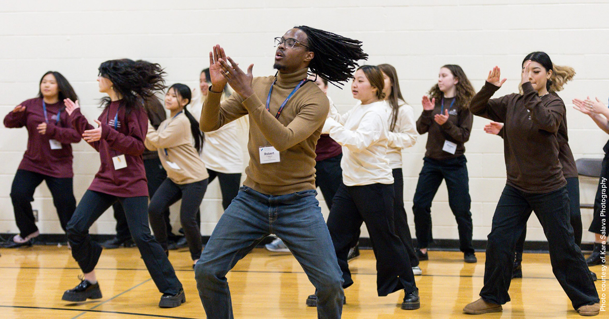 Teenage participants in the Urban Choir Festival wearing long sleeved shirts of different colors and black pants while clapping and dancing led by an instructor with facial hair and glasses wearing a tan turtleneck and jeans clapping and dancing. Photo Credit: Kara Salava Photography.