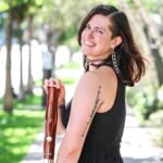 Image of person wearing a black sleeveless dress with dark hair looking over their shoulder and smiling while holding a bassoon in a sunlight sidewalk with trees in the background.