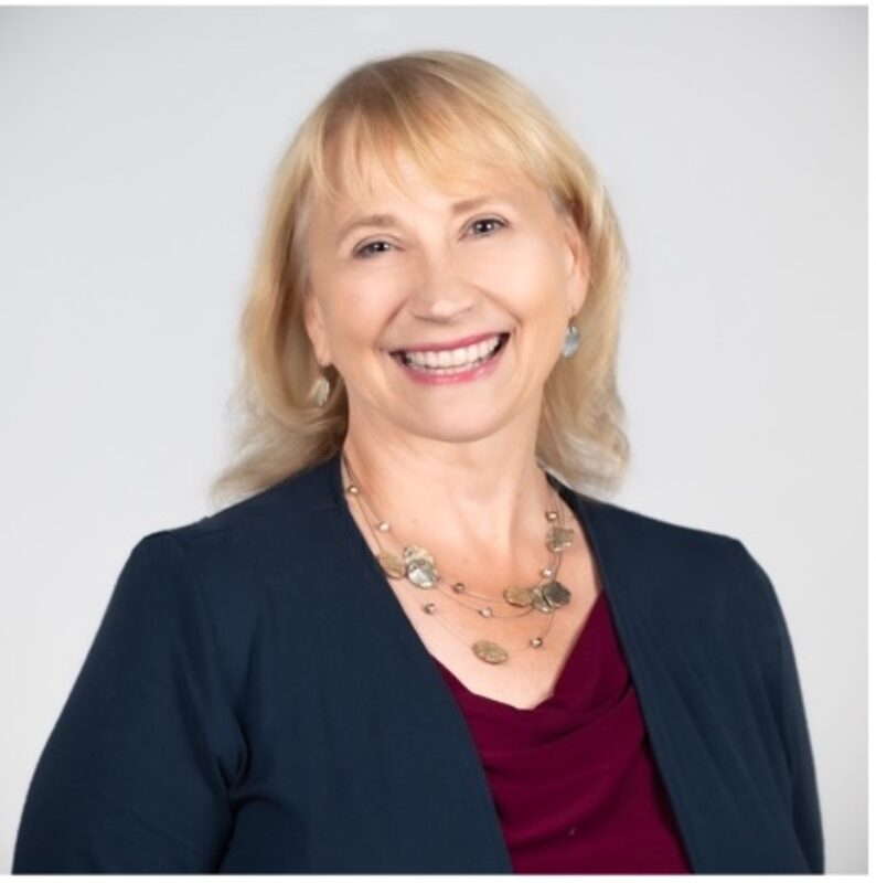 Person with blond shoulder length hair wearing earrings and a necklace with a maroon shirt and navy blue blazer.